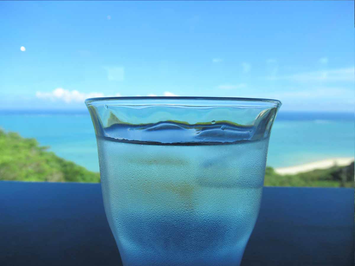 沖縄南部　カフェやぶさちの窓辺に見える青い海をお水のコップを通してみる写真